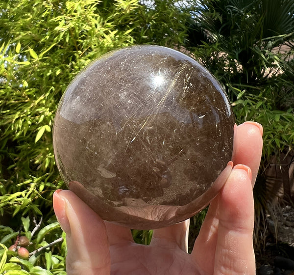 Image of a hand holding a rare golden rutilated smoky quartz crystal sphere. 